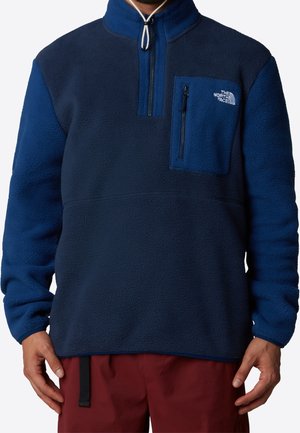 Man draagt een tweekleurige marineblauwe fleece trui met een ritssluiting op de borstzak en het The North Face-logo aan de linkerbovenkant.