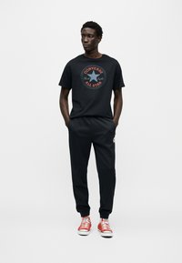 Μαύρο βαμβακερό μπλουζάκι με κόκκινο και μπλε κυκλικό λογότυπο, συνδυασμένο με μαύρο παντελόνι jogging και κόκκινα αθλητικά παπούτσια με λευκές σόλες.