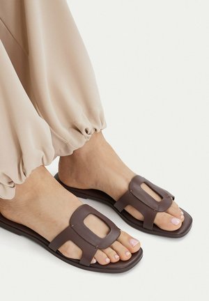 Piedi con pantaloni beige e sandali aperti color marrone scuro con tagli rettangolari, mostrati su uno sfondo bianco.