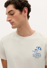 Hellbeige Baumwoll-T-Shirt mit einem blauen Grafikdruck eines Hundes unter einem Schirm, versehen mit dem Text „DER SOMMER ENDEN IM SÜDEN NIE.“