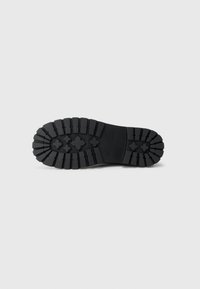 Suela de zapato negra con surcos profundos y tacos en forma de cruz para mayor agarre, mostrada plana contra un fondo gris claro.
