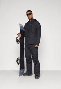 Fekete vízálló dzseki elől cipzárral, fekete hó nadrággal párosítva. A snowboard sötét felülettel rendelkezik, mintás teteje van.