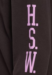 Tamna tkanina vertikalno otisnuta s ružičastim slovima "H. S. W.", prikazujući detaljnu teksturu materijala.