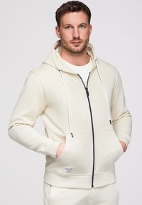 Homme portant un sweat à capuche crème zippé, les mains dans les poches, regardant devant avec une expression neutre sur un fond uni.