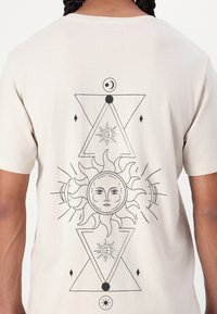 Tricou bej cu mânecă scurtă, prezentând un design cu artă liniară neagră a unui soare și forme geometrice pe spate, cu o textură moale din bumbac.