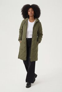 Veste longue matelassée vert olive avec capuche, dotée de poches latérales et de poignets côtelés, associée à un t-shirt blanc et un pantalon noir à jambes larges.
