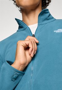 Teal fleece zip-up jack met hoge kraag, voorzien van een zichtbare rits en logodetail op de borst. Zachte textuur en verstelbare manchetten.