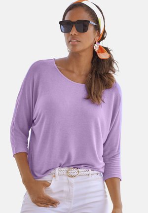 Lavendelfarbenes Langarmoberteil mit weitem Ausschnitt, kombiniert mit weißen Hosen und einem geflochtenen Gürtel. Die Accessoires umfassen Sonnenbrillen und ein farbenfrohes Stirnband.