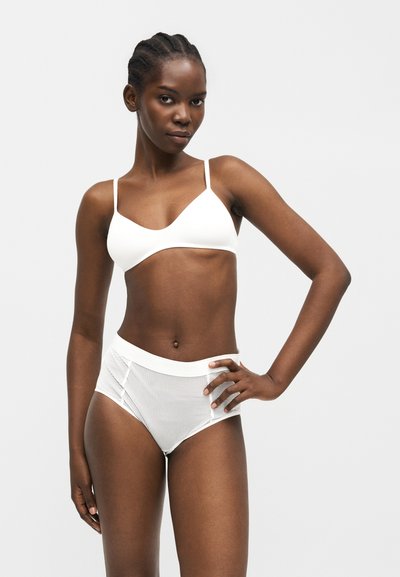 Γυναίκα με πλεγμένα μαλλιά που φοράει λευκό μπραλέτα και λευκά και γκρι ριγέ εσώρουχα με ψηλή μέση, στέκεται με το ένα χέρι στη μέση της.