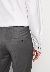 Grau maßgeschneiderte Hose aus strukturiertem Stoff, mit einer Gesäßtasche mit einem schwarzen Knopf und einem weißen Hemd mit blauen Akzenten an den Manschetten.