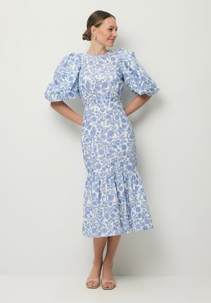 Vrouw die een blauw-witte midi-jurk met bloemenprint draagt, met opgeblazen mouwen en een gerimpelde zoom, staand tegen een effen lichte achtergrond.