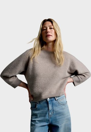 Blonde vrouw staat met handen in de zij, draagt een losse grijze trui en een hoog getailleerde blauwe spijkerbroek tegen een effen achtergrond.