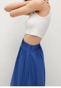 Persona vestita con un top corto senza maniche bianco e una gonna plissettata blu a vita alta, in piedi di lato con le braccia sollevate dietro la testa.