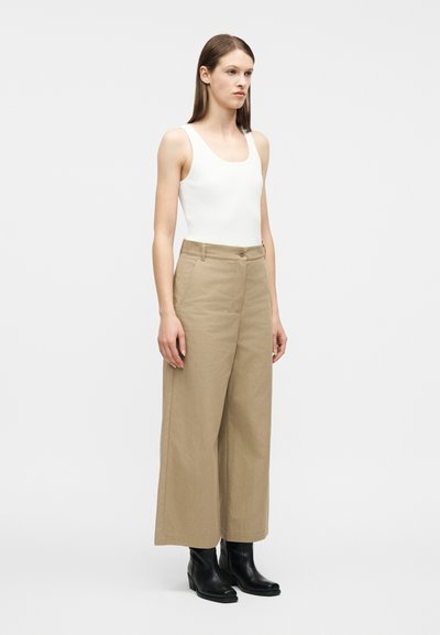 Γυναίκα που στέκεται φορώντας λευκό αμάνικο ριμπ τοπ, μπεζ παντελόνι με φαρδιά μπατζάκια και μαύρα δερμάτινα μποτάκια στον αστράγαλο πάνω σε απλό λευκό φόντο.