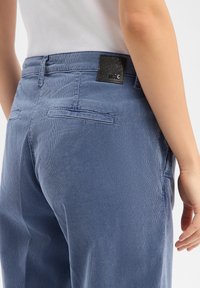 Blue corduroy trousers with a textured finish, featuring a black leather brand patch and two back pockets, shown from the side and back.