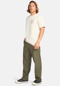 T-shirt en coton beige clair pour homme avec manches courtes et design graphique sur le devant, associé à un pantalon vert olive et des baskets noires.
