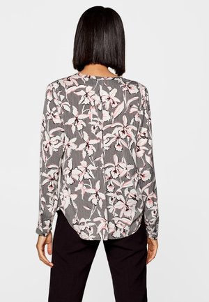 Vrouw met recht zwart haar, gekleed in een blouse met lange mouwen in wit, roze en zwart bloemenpatroon en zwarte broek, gezien van achteren.