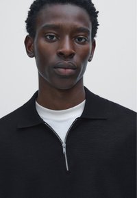 Young man with short curly hair wearing a black collared sweater with a half-zip over a white crew neck shirt against a plain background.