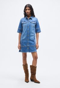 Φόρεμα από τζιν ύφασμα με κοντά μανίκια, κλείσιμο με κουμπιά στο μπροστινό μέρος, τσέπες στο στήθος και αντίθετες ραφές· συνδυασμένο με καφέ καουμπόικες μπότες από καστόρι.