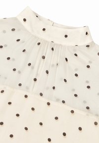 Herkullinen pusero, jossa on korkea kaulus, kermaisen värinen ja musta-beigeiä täpliä. Sisältää poimutus- ja nappikiinnityksen takana.