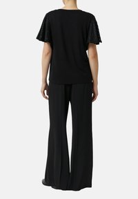 Person trägt schwarze, weit geschnittene Hosen und ein schwarzes Kurzarmoberteil mit strukturierten, ausgestellten Ärmeln, steht mit dem Rücken zur Kamera vor einem weißen Hintergrund.