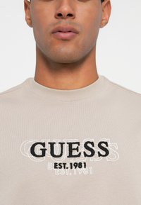 Primer plano de una persona que lleva una camisa de color beige claro con "GUESS EST. 1981" bordado en negro y blanco en el pecho.
