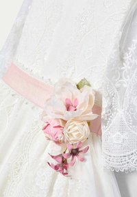 Abito bianco con dettagli in pizzo intricati, una morbida fascia rosa e un insieme di fiori di stoffa bianchi, rosa e lavanda.