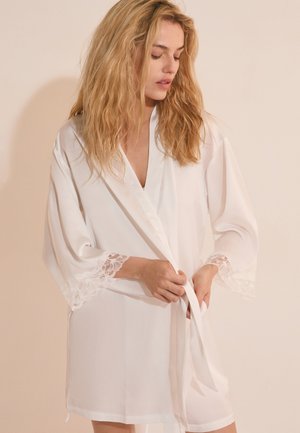 Femme aux longs cheveux blonds portant une robe blanche bordée de dentelle, attachant la ceinture à la taille, debout devant un fond neutre.