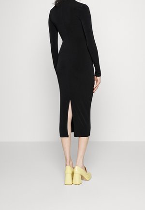 Robe midi noire à manches longues avec une fente au dos, silhouette ajustée et texture douce. Une paire de chaussures à talons carrés jaunes est visible en bas.