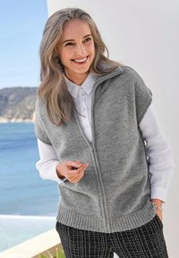 Un gilet tricoté gris clair à manches courtes, avec fermeture éclair à l'avant et ourlet côtelé, porté par-dessus une chemise blanche à col, associé à un pantalon noir à motifs.