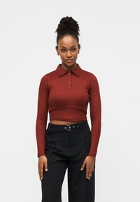 Μακρυμάνικο polo κοντομάνικο σε σκούρο καφέ ριπ ύφασμα με πέντε κουμπιά και γιακά, συνδυασμένο με ψηλόμεσο μαύρο παντελόνι.