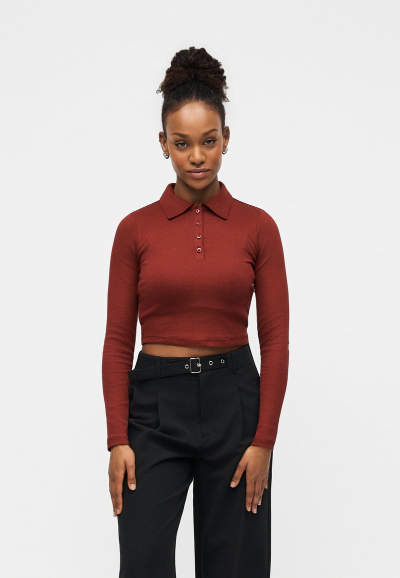 Μακρυμάνικο polo κοντομάνικο σε σκούρο καφέ ριπ ύφασμα με πέντε κουμπιά και γιακά, συνδυασμένο με ψηλόμεσο μαύρο παντελόνι.