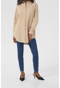 Chemise beige à boutons avec une poche poitrine, manches longues et coupe décontractée, associée à un jean skinny bleu et des mules noires à talons.