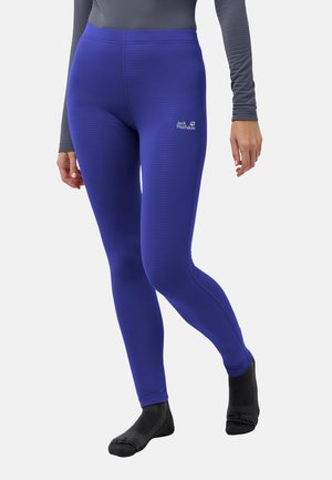 Persona che indossa leggings blu Jack Wolfskin e calzini neri, in piedi con una gamba leggermente sollevata, su uno sfondo chiaro uniforme.