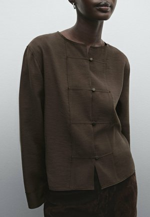 Persona che indossa una maglia a maniche lunghe marrone scuro con dettagli visibili di bottoni e scollo rotondo, in piedi davanti a uno sfondo chiaro e semplice.