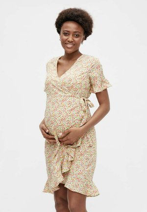Pregnant woman in a floral wrap dress gently holding her belly and smiling against a plain white background.