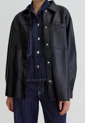Person trägt eine schwarze, übergroße Lederjacke über einem dunkelblauen Nadelstreifen-Denimhemd und Hose, steht vor neutralem Hintergrund.