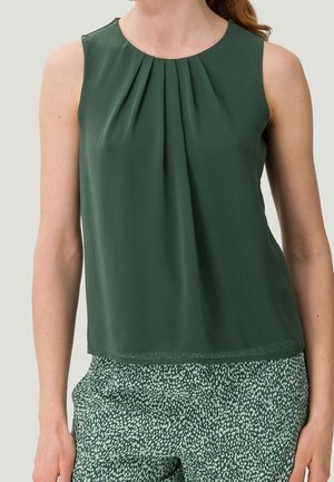 Femme portant un haut sans manches vert foncé avec des plis au niveau de l'encolure et un pantalon vert à motifs sur fond uni.