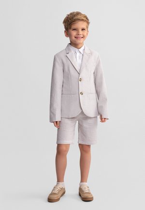 Uśmiechnięty chłopiec, ubrany w jasny, teksturowany blezer i dopasowane szorty, białą koszulę oraz beżowe trampki, stojący na jednolitym tle.