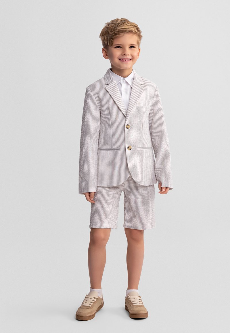 Jeune garçon souriant, portant un blazer texturé de couleur claire et un short assorti, avec une chemise blanche et des baskets beiges, debout devant un fond uni.