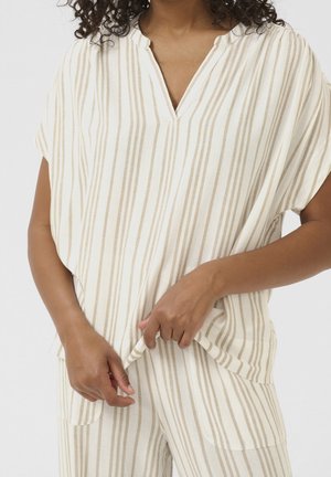 Kvinde iført løs beige og hvide vertikale stribede kortærmede top og matchende bukser, med hænderne let holdende i toppen af tøjet.