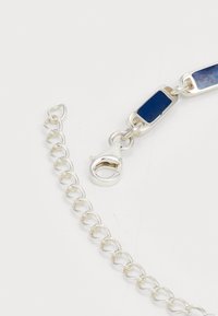 Ezüst nyaklánc kapocs kék téglalap alakú láncszemekkel és egy hosszabbító lánccal fehér háttér előtt.