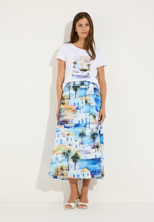 Woman wearing white t-shirt with beach print and long skirt featuring blue, white, and green Mediterranean seaside landscape, standing against plain wall.