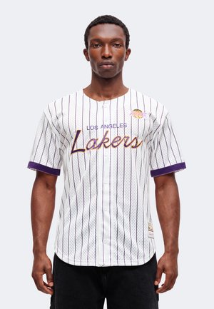 Jeune homme portant un maillot de baseball blanc des Los Angeles Lakers avec des fines rayures violettes et des poignets violets, debout devant un fond uni.