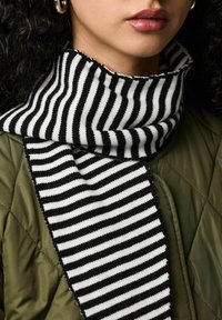 Écharpe tricotée avec des rayures alternées noires et blanches, présentant une texture côtelée. Portée sur une veste matelassée vert olive.