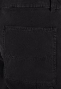 Gros plan d'une poche arrière en tissu noir sur un pantalon avec des coutures visibles et une boucle de ceinture au-dessus.