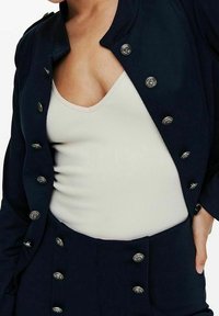 Blazer bleu marine avec détails de boutons argentés, associé à un débardeur blanc côtelé. Le blazer présente un col arrondi et une coupe ajustée.