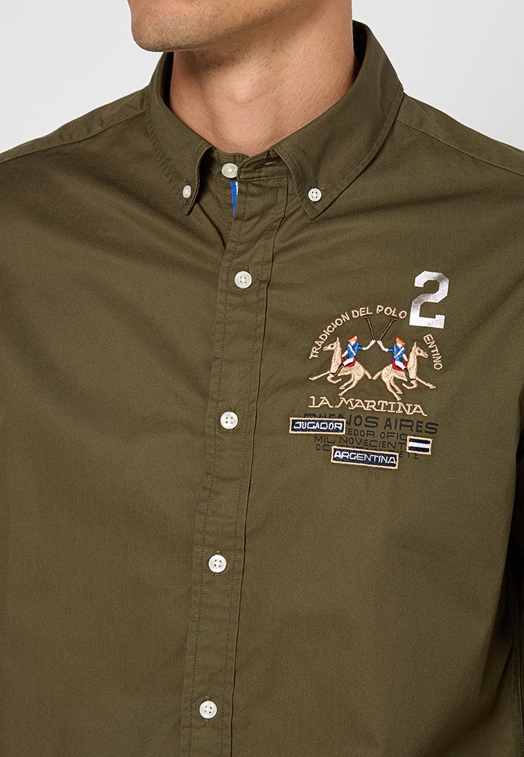 Camicia verde oliva a maniche lunghe in cotone. Presenta un motivo ricamato di polo, toppe e un colletto strutturato. Bottoni bianchi e dettagli blu.