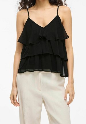 Kobieta w czarnej, warstwowej bluzce bez rękawów z cienkimi ramiączkami, w połączeniu z luźnymi białymi spodniami na jednolitym tle.