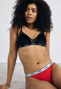 Woman with curly hair wearing black velvet bralette and red underwear with white elastic bands branded "HUGO," sitting on a light-colored carpet.